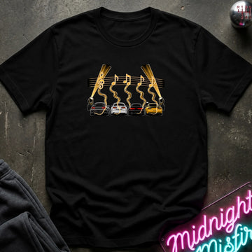 A black short-sleeve t-shirt laid flat on a dark textured surface, featuring a graphic print of four cars (red, white, gray, yellow) with gold wavy lines and musical notes above them, flanked by two  lights.