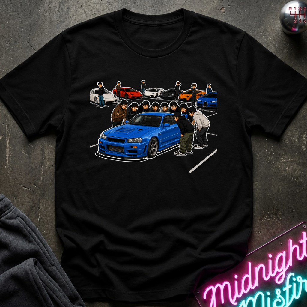 A black short-sleeve graphic t-shirt laid flat on a dark textured surface. The shirt features a detailed graphic print of a blue sports car with several anime-style characters standing around it, and smaller cars in the background.