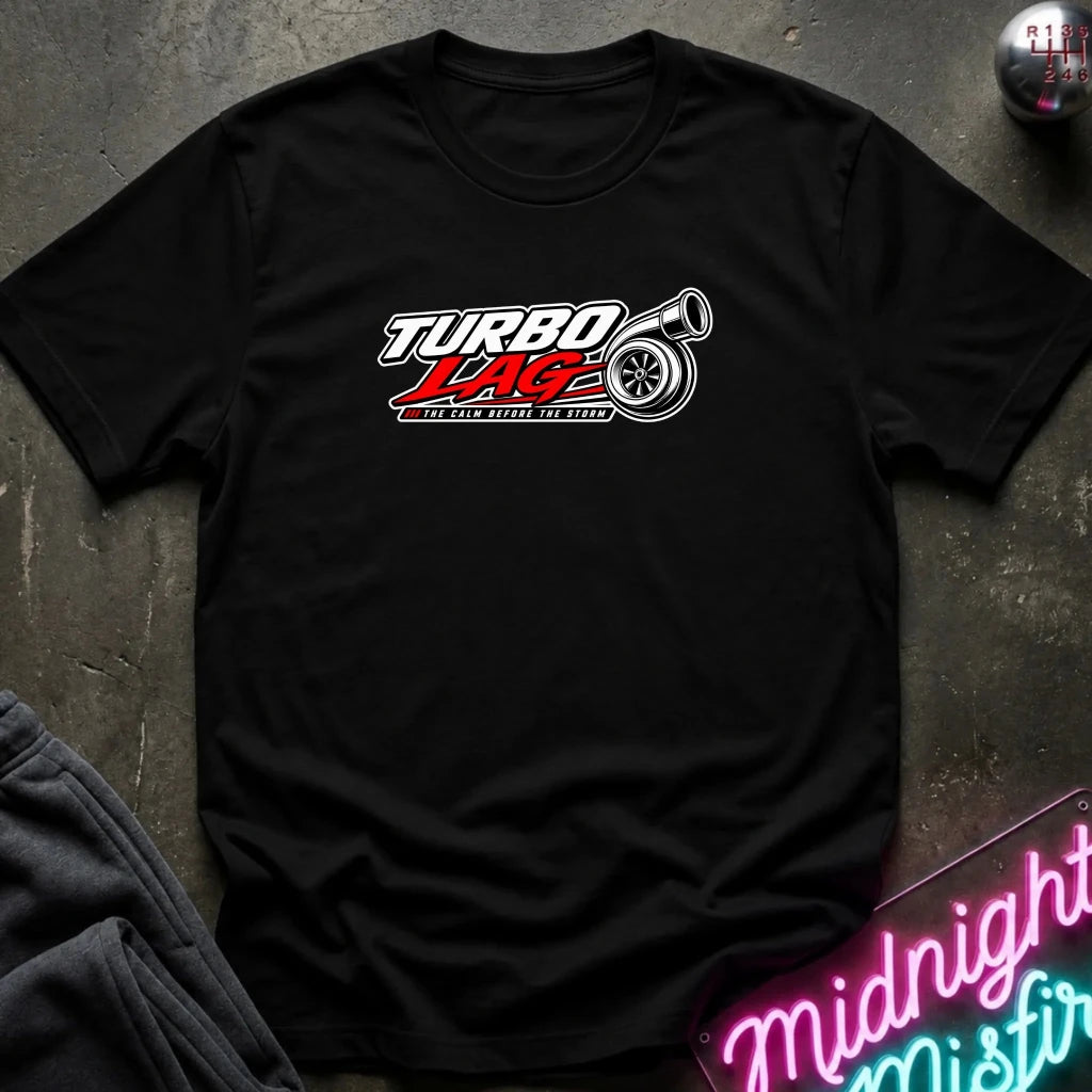 A black short-sleeve t-shirt laid flat on a dark textured surface, featuring a 'Turbo Lagz' graphic on the chest with a stylized tire and red lettering. A neon sign and folded dark fabric are partially visible in the corners.