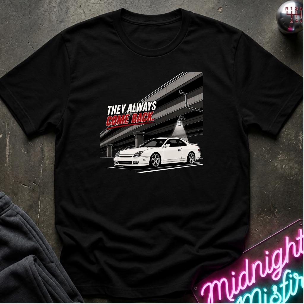 A black short-sleeve graphic t-shirt laid flat on a dark textured surface. The graphic features a white sports car under a overpass with the text 'THEY ALWAYS COME BACK' in white and red.