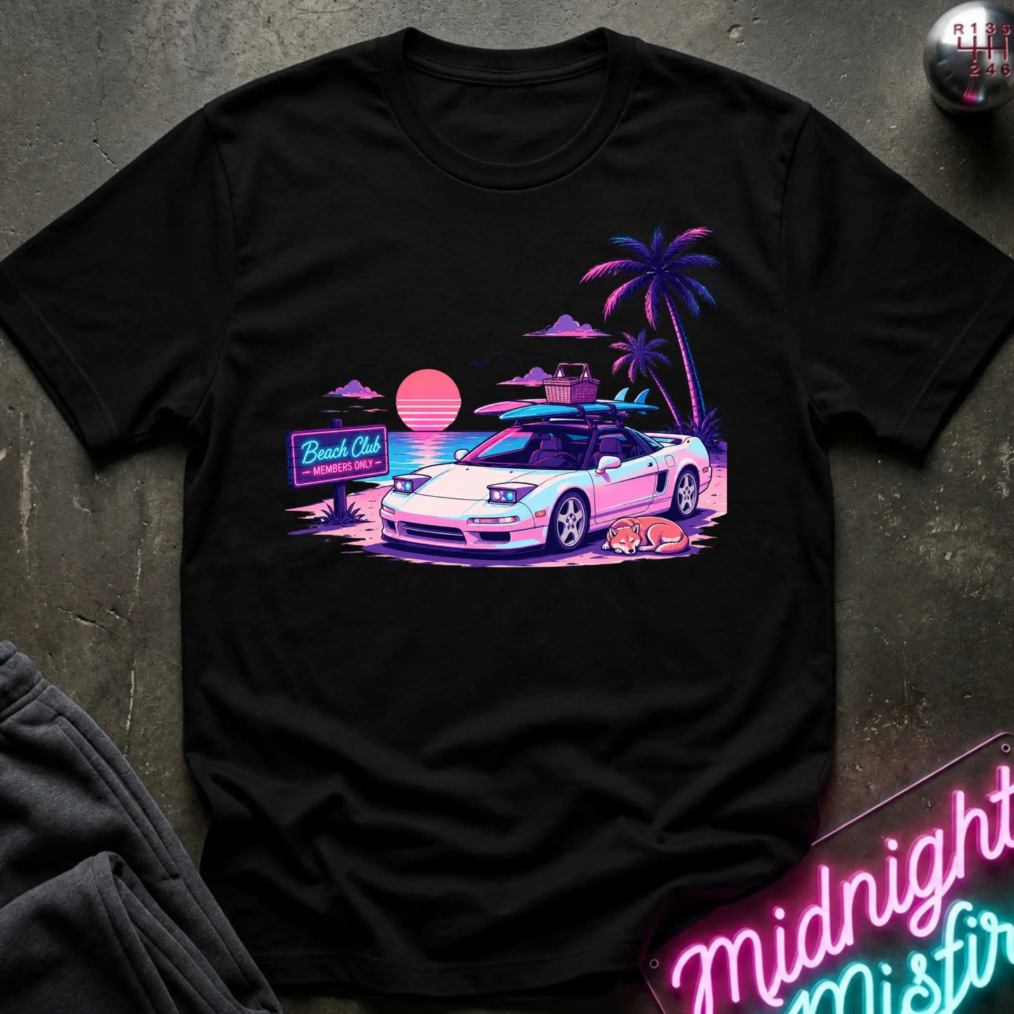 A black short-sleeve graphic t-shirt laid flat on a dark textured surface. The graphic features a retro-style illustration of a sports car with surfboards on its roof, next to palm trees, a sunset, and a 'Beach Club' sign.