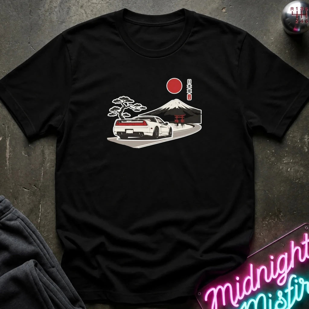 A black short-sleeve graphic t-shirt laid flat on a dark textured surface. The graphic shows a white sports car driving toward a Japanese-style mountain and red sun, with a red circle and vertical text.