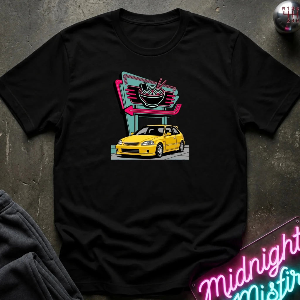 A black short-sleeve t-shirt laid flat on a dark textured surface, featuring a retro-style graphic of a yellow hatchback car parked in front of a neon ramen shop sign. 