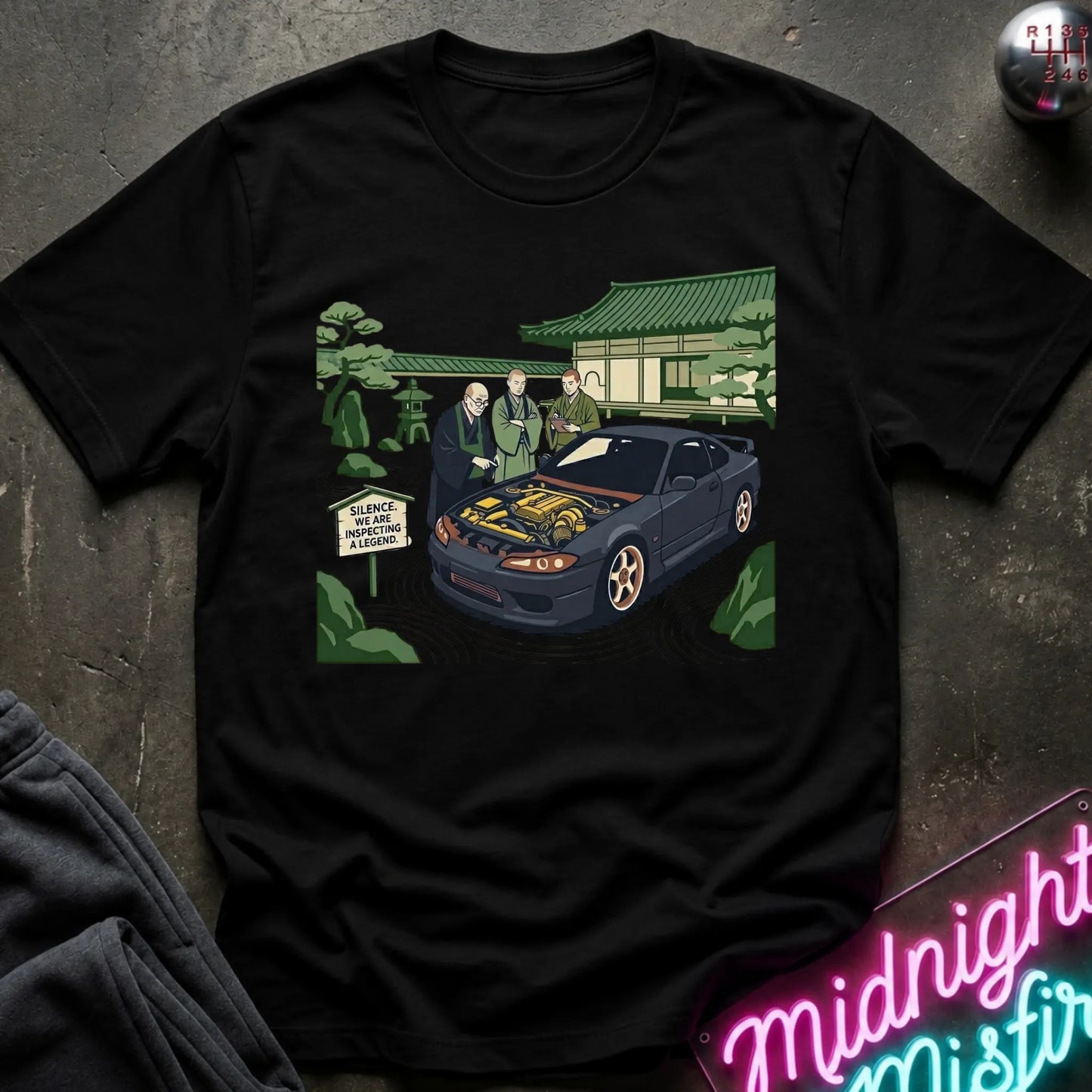 Black short-sleeve graphic t-shirt laid flat on a dark textured surface. The graphic shows a black sports car with its hood open, surrounded by two men in suits, a Japanese-style building, and green trees.