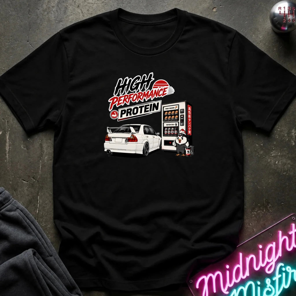 A black short-sleeve graphic t-shirt laid flat on a dark textured surface. The graphic features a white car, a vending machine, and the text 'High Performance Protein' in stylized red, white, and black lettering.