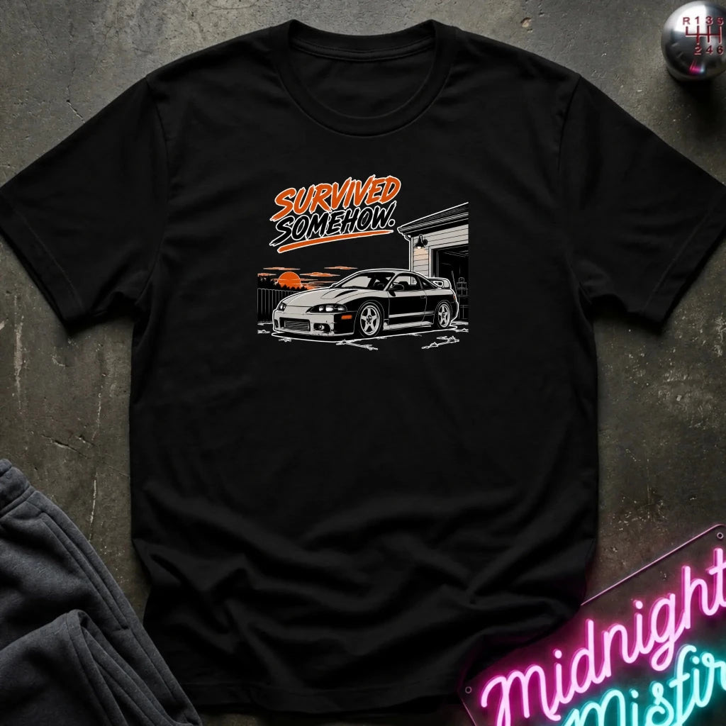 A black short-sleeve graphic t-shirt laid flat on a dark textured surface. The graphic features the words 'SURVIVED SOMEHOW' in orange and white above a white sports car in front of a garage with an orange sunset.
