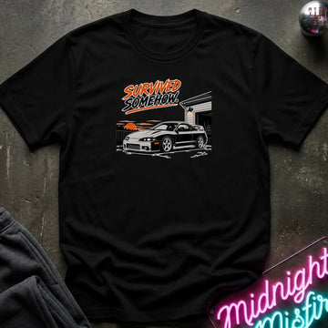 A black short-sleeve graphic t-shirt laid flat on a dark textured surface. The graphic features the words 'SURVIVED SOMEHOW' in orange and white above a white sports car in front of a garage with an orange sunset.