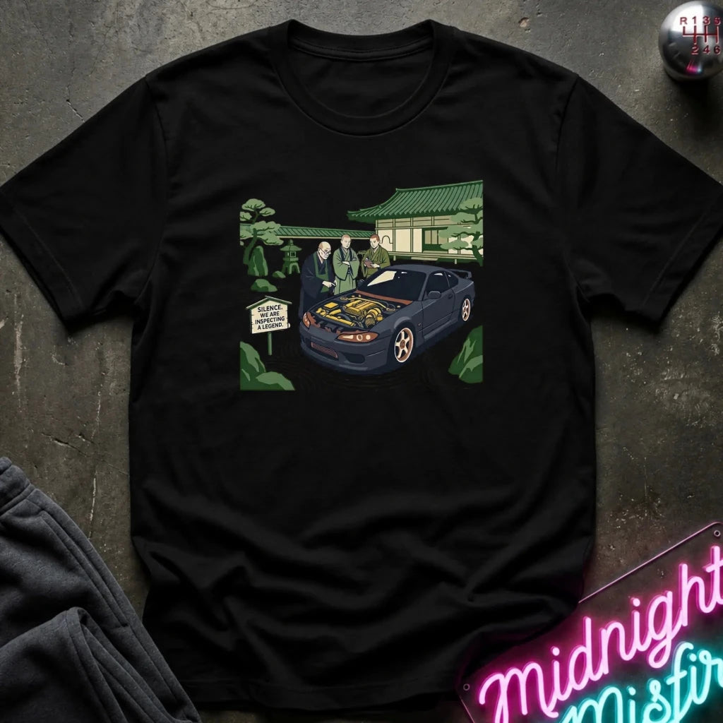 A black short-sleeve graphic t-shirt laid flat on a dark textured surface. The graphic shows a dark blue sports car with its hood open, with two men standing beside it in front of a building with a green roof.