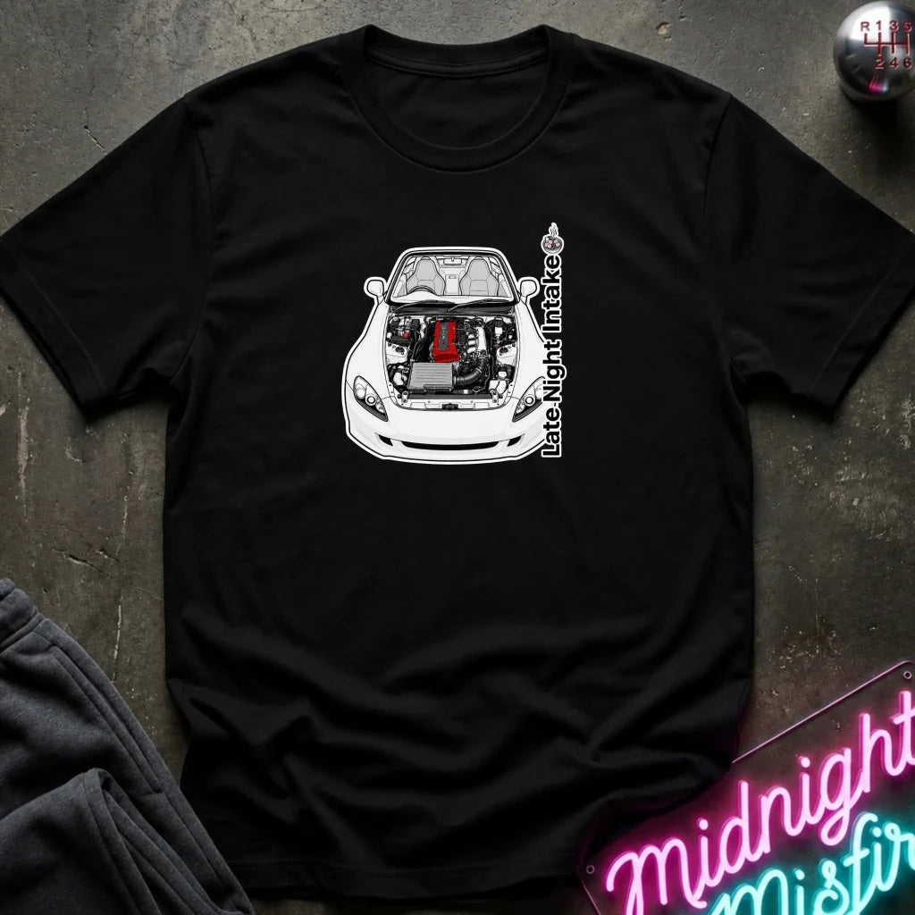 A black short-sleeve t-shirt laid flat on a dark textured surface, featuring a graphic of a white sports car with its hood open, revealing a red engine component, alongside the vertical text 'Late Night Intake'.