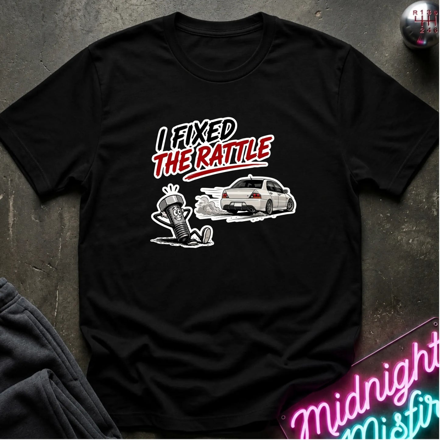 A black short-sleeve graphic t-shirt laid flat on a dark textured surface. The graphic features the text 'I FIXED THE RATTLE' in white and red, above a white car and a cartoon character sitting on springs.