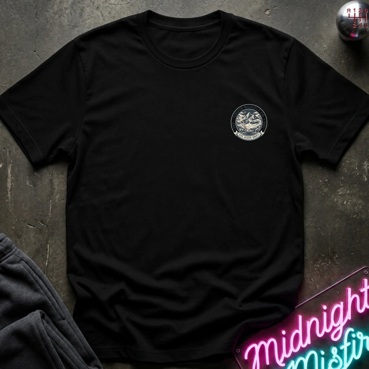 Black crewneck t-shirt with a circular crest printed on the left chest, styled flat on a dark textured surface with a neon sign and dark pants in the background.