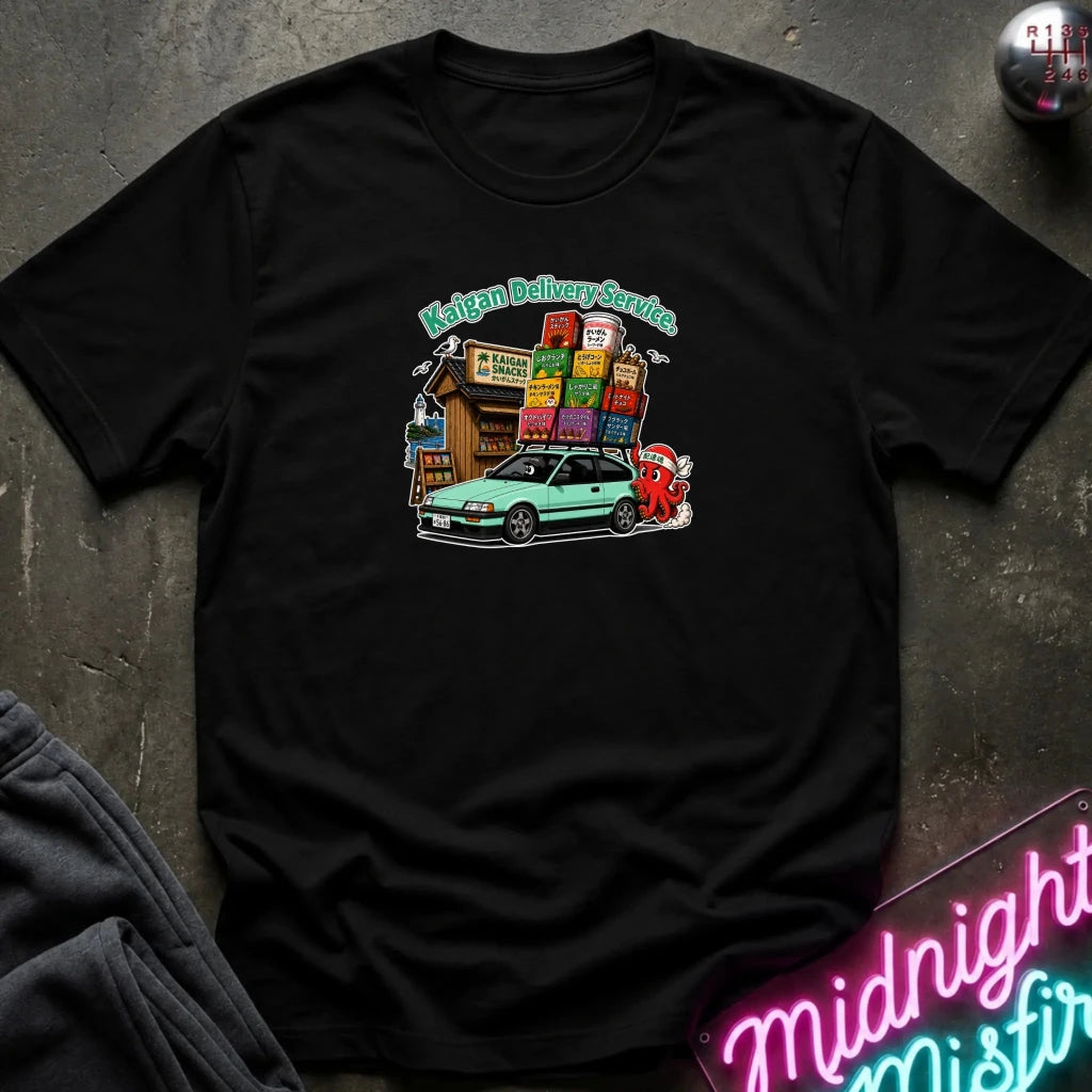 A black graphic t-shirt laid flat on a dark textured surface. The graphic features a light green car, and a small building with colorful signs under the text 'Kaigan Delivery Service'.
