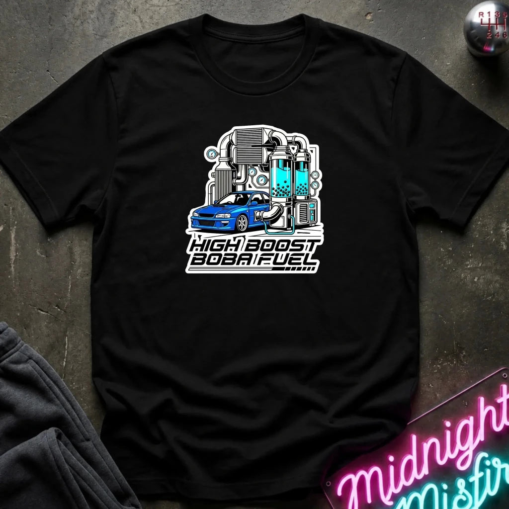 A black short-sleeve graphic t-shirt laid flat on a dark textured surface. The shirt features a white, gray, and blue illustration of a sports car connected to industrial pipes and tanks, with the text 'HIGH BOOST BOBAFUEL' printed below.