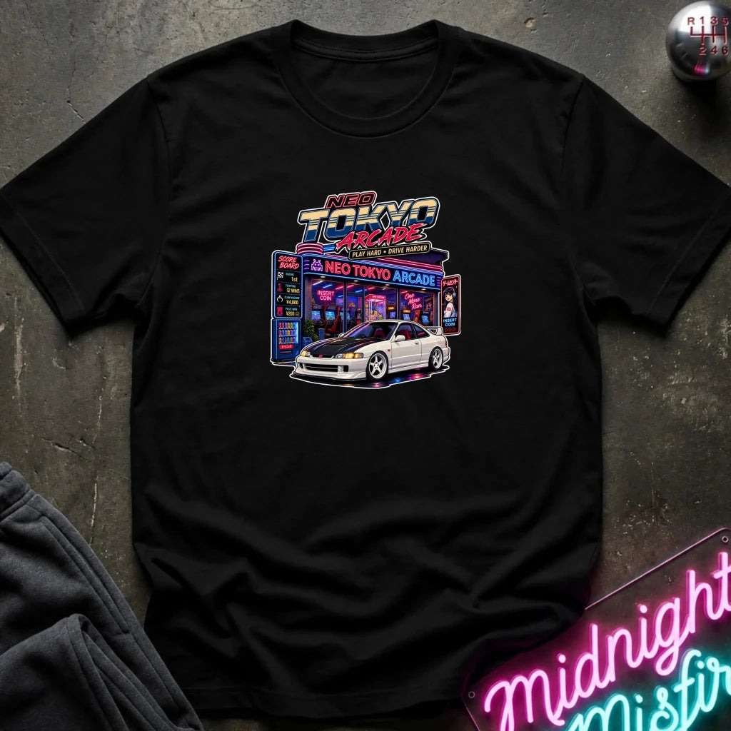 A black short-sleeve crewneck t-shirt featuring a 'Neo Tokyo Arcade' graphic with a white sports car, laid flat on a dark textured surface with a neon sign and sweatpants as props.