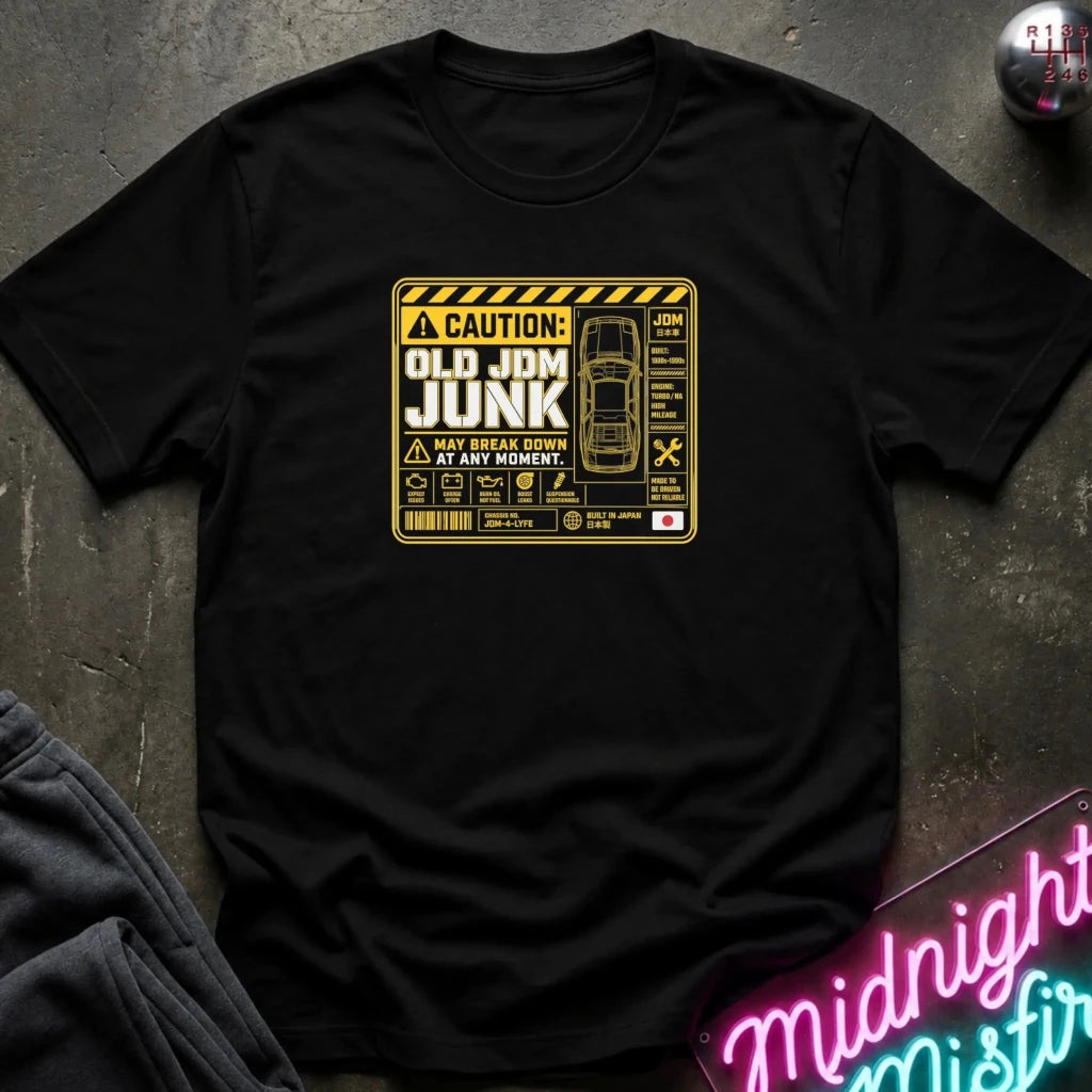 A black short-sleeve t-shirt laid flat on a dark textured surface, featuring a yellow and black caution-style graphic on the chest that reads 'OLD JDM JUNK' with a car illustration and warning symbols.