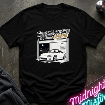 A black short-sleeve graphic t-shirt laid flat on a dark textured surface. The graphic shows a white convertible car parked in front of a garage with the text 'THEY DON'T MAKE THEM LIKE THIS ANY MORE' in stylized lettering.