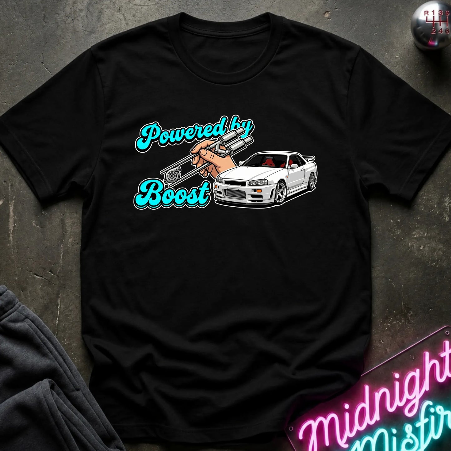 A black graphic t-shirt laid flat on a dark textured surface. The graphic features the text 'Powered by Boost' in a teal cursive font, with a hand holding a silver turbocharger, alongside a white sports car.