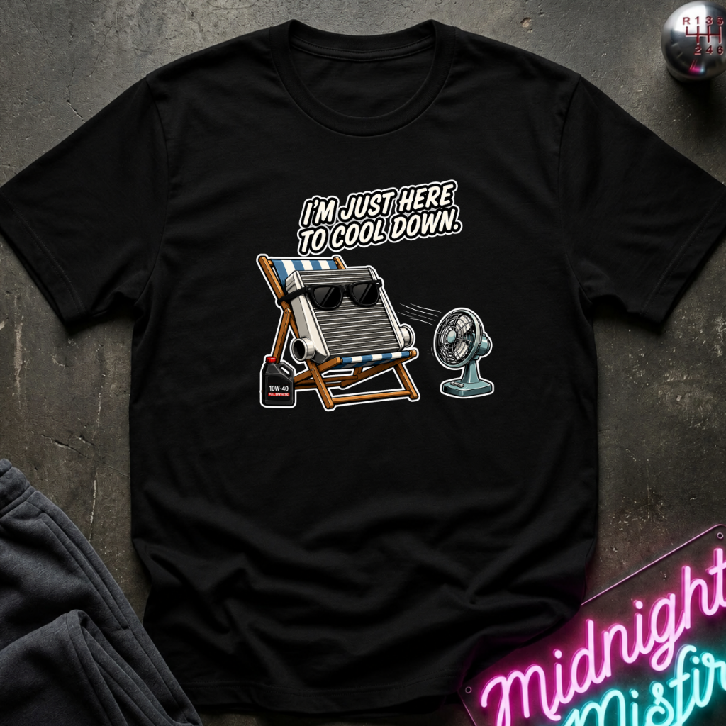 A black short-sleeve graphic t-shirt laid flat on a dark textured surface. The graphic features a striped beach chair and radiator wearing sunglasses with the text 'I'M JUST HERE TO COOL DOWN.
