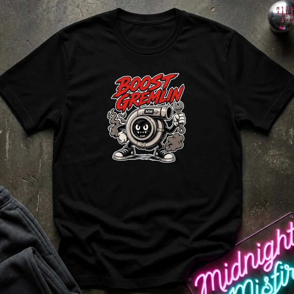 A black short-sleeve graphic T-shirt laid flat on a dark textured surface. The shirt features a cartoon character with a mustache and goggles, with the words 'BOOST GREMLIN' in red and white above it.