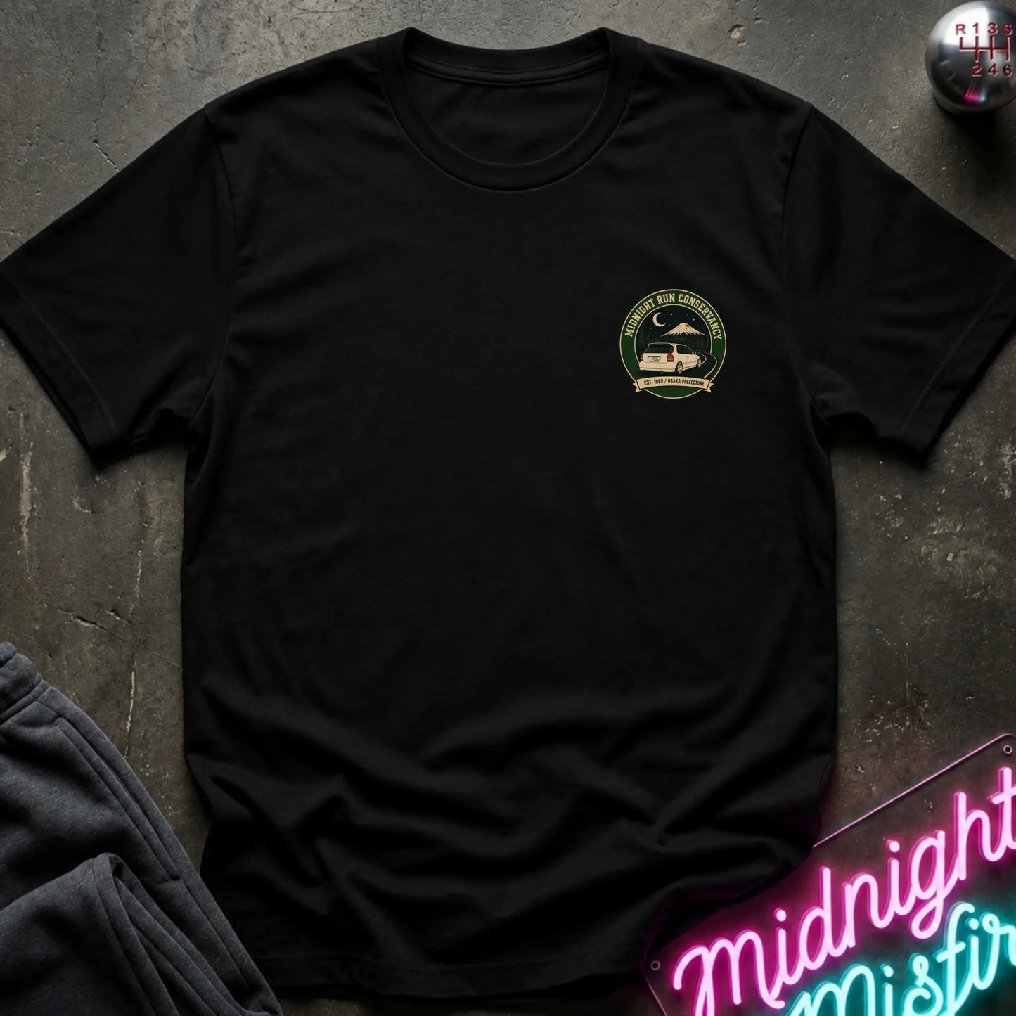 A black short-sleeve crewneck t-shirt with a circular green and black chest graphic, styled flat-lay on a dark textured surface with a neon sign and dark pants nearby.