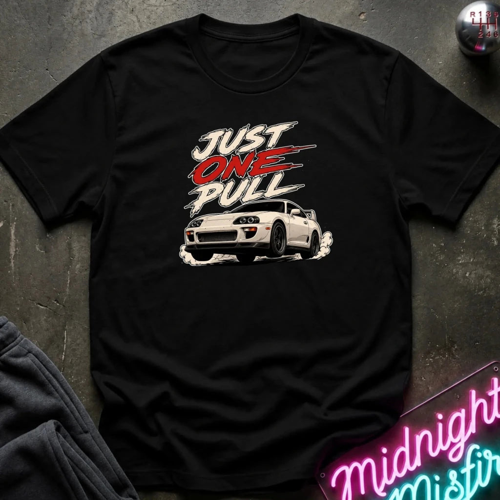 A black short-sleeve graphic t-shirt laid flat on a dark textured surface. The graphic features the text 'JUST ONE PULL' in stylized white and red lettering above a white sports car with smoke effects.