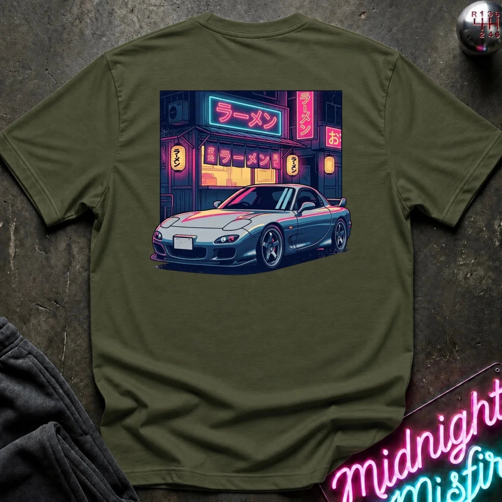 An olive green t-shirt laid flat on a dark textured surface, featuring a colorful graphic print of a blue sports car parked in front of a neon-lit ramen shop.