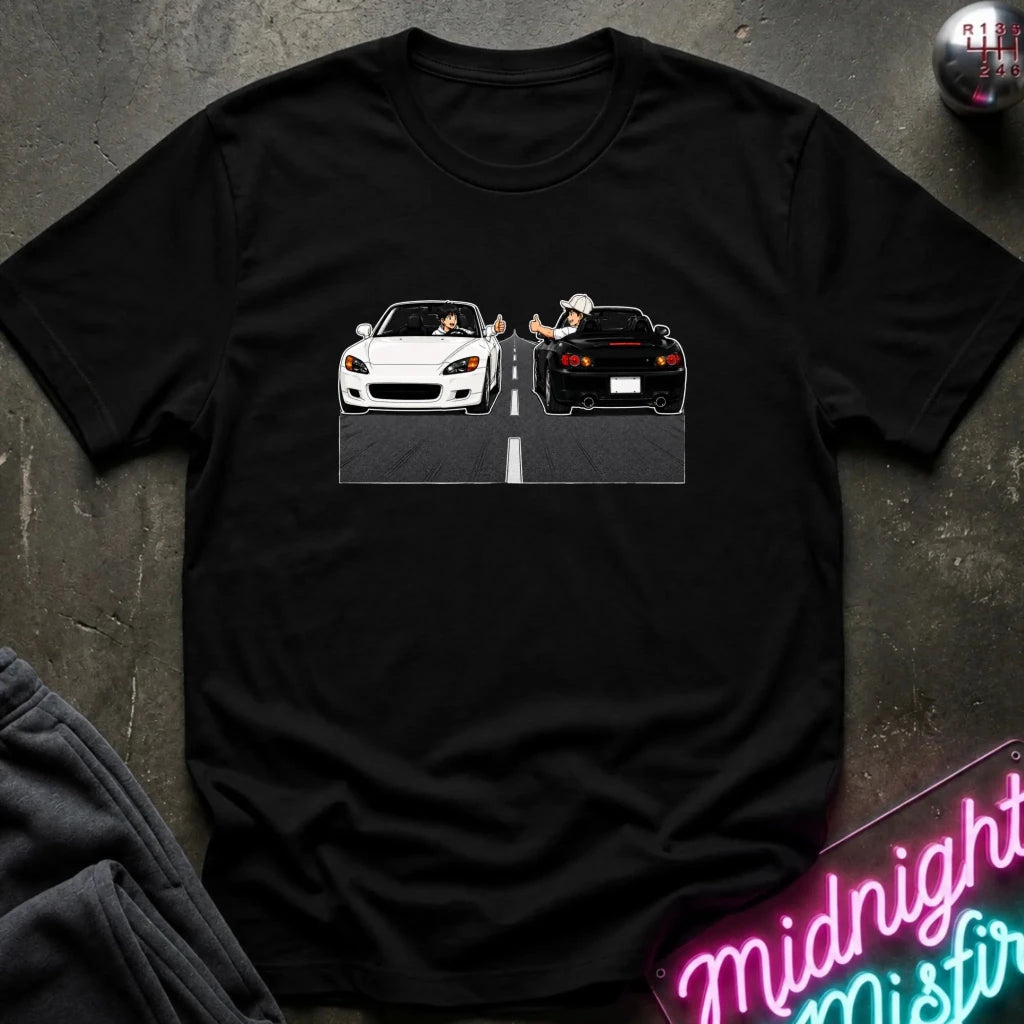 A black short-sleeve t-shirt laid flat on a dark textured surface, featuring a graphic print of a white convertible sports car facing a black sports car in a split scene. Styled flat-lay props include a dark sweatshirt in the lower-left corner and a neon sign in the lower-right.