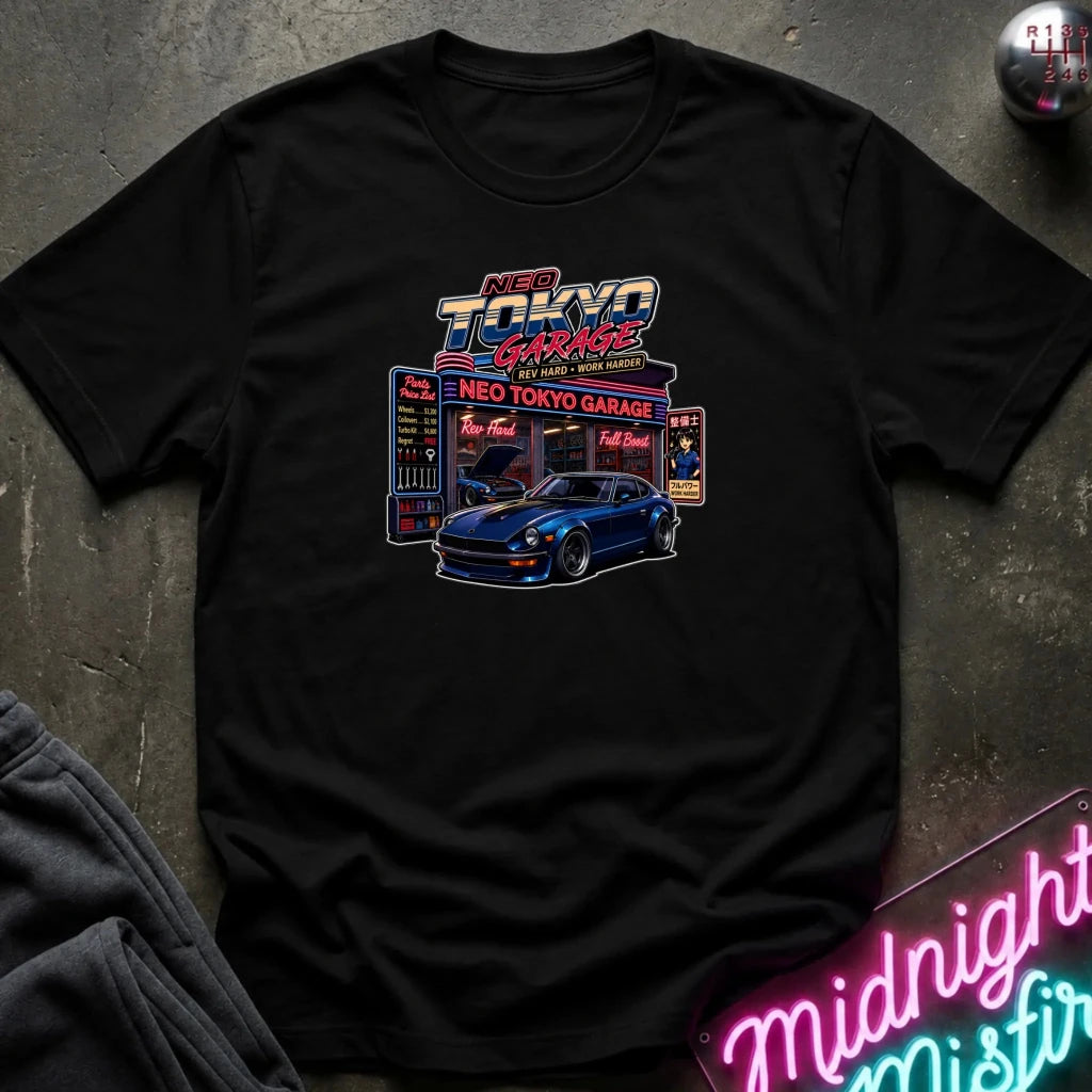 A black short-sleeve graphic T-shirt laid flat on a dark textured surface. The shirt features a colorful retro-style print reading 'Neo Tokyo Garage' with a blue sports car and neon sign elements.