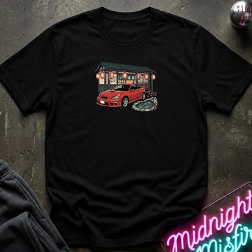 A black short-sleeve t-shirt laid flat on a dark textured surface, featuring a graphic print of a red sports car parked in front of a Japanese-style building with lanterns. 
