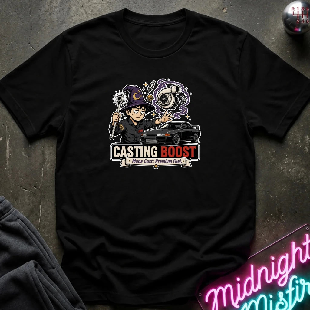 A black short-sleeve graphic t-shirt laid flat on a dark textured surface. The graphic features a cartoon character holding a sign that reads 'CASTING BOOST', a black car, and a purple smoke effect.