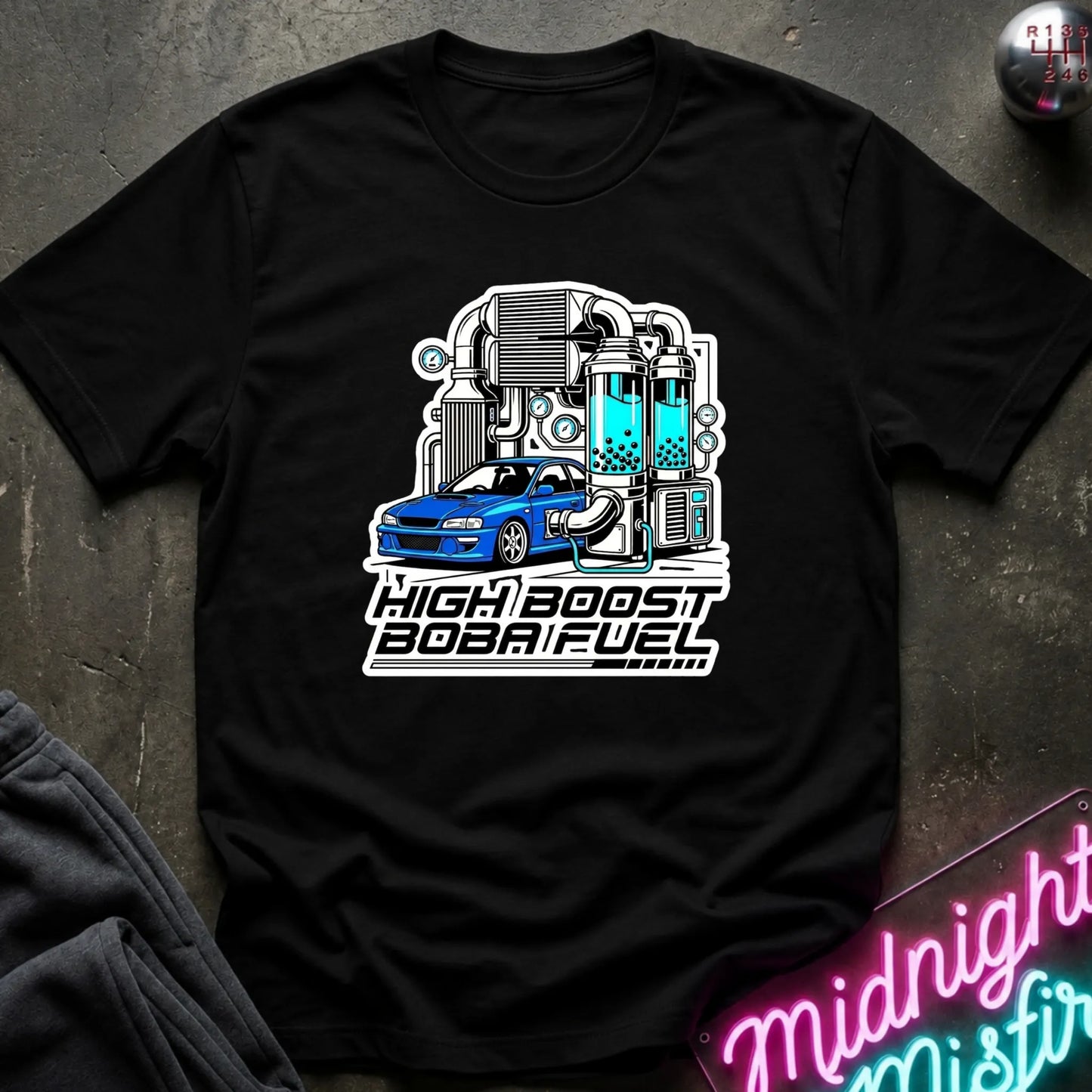 A black short-sleeve graphic t-shirt laid flat on a dark textured surface. The shirt features a stylized graphic of a blue car connected to industrial machinery with cyan liquid, above the text 'HIGH BOOST BOBAT FUEL'.