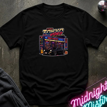 A black short-sleeve crewneck t-shirt featuring a 'Neo Tokyo video' graphic laid flat on a dark textured surface with a neon sign and sweatpants as props.