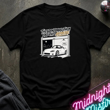 A black short-sleeve graphic t-shirt laid flat on a dark textured surface. The graphic features a white car inside a garage with stylized text above it.