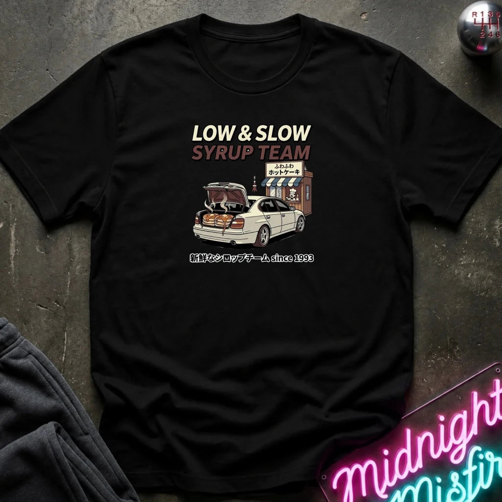A black short-sleeve graphic T-shirt laid flat on a dark textured surface. The shirt features a printed design with the text 'LOW & SLOW SYRUP TEAM' above an illustration of a white car with its trunk open in front of a small shop, with Japanese text below.