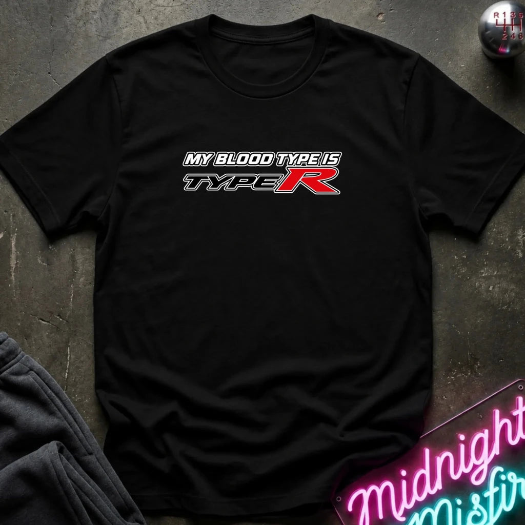 A black short-sleeve crewneck t-shirt featuring a 'MY BLOOD TYPE IS TYPE R' graphic on the chest, laid flat on a dark textured surface with a neon sign and clothing in the background.