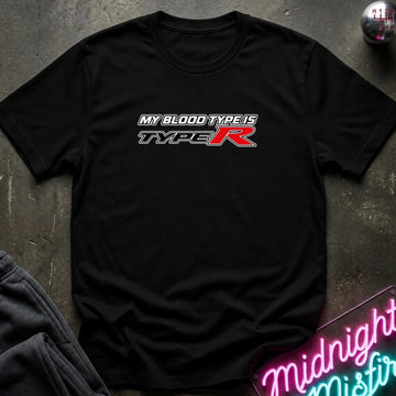 A black short-sleeve crewneck t-shirt featuring a 'MY BLOOD TYPE IS TYPE R' graphic on the chest, laid flat on a dark textured surface with a neon sign and clothing in the background.
