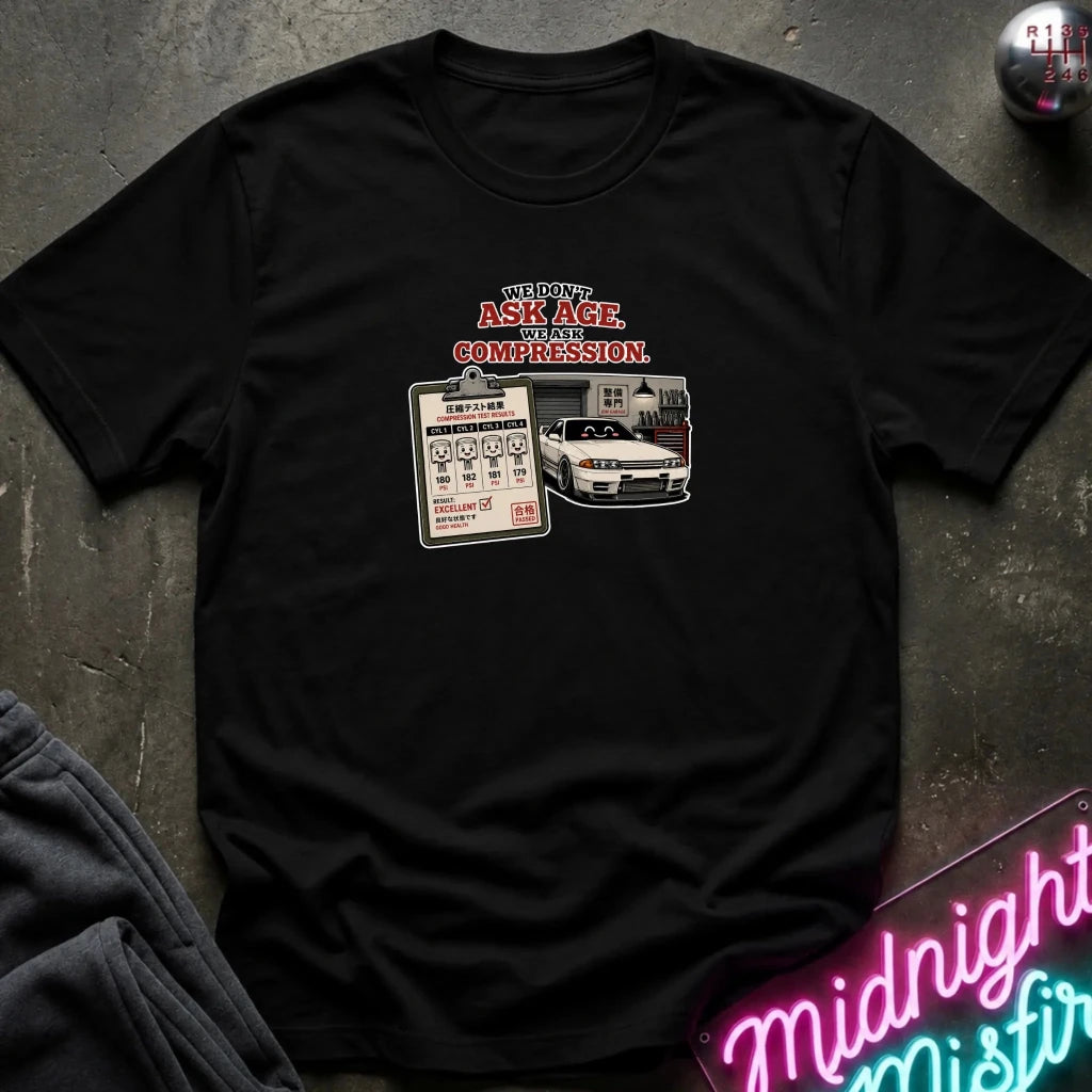 A black short-sleeve graphic t-shirt laid flat on a dark textured surface. The graphic features the text 'WE DON'T ASK AGE, WE ASK COMPRESSION.