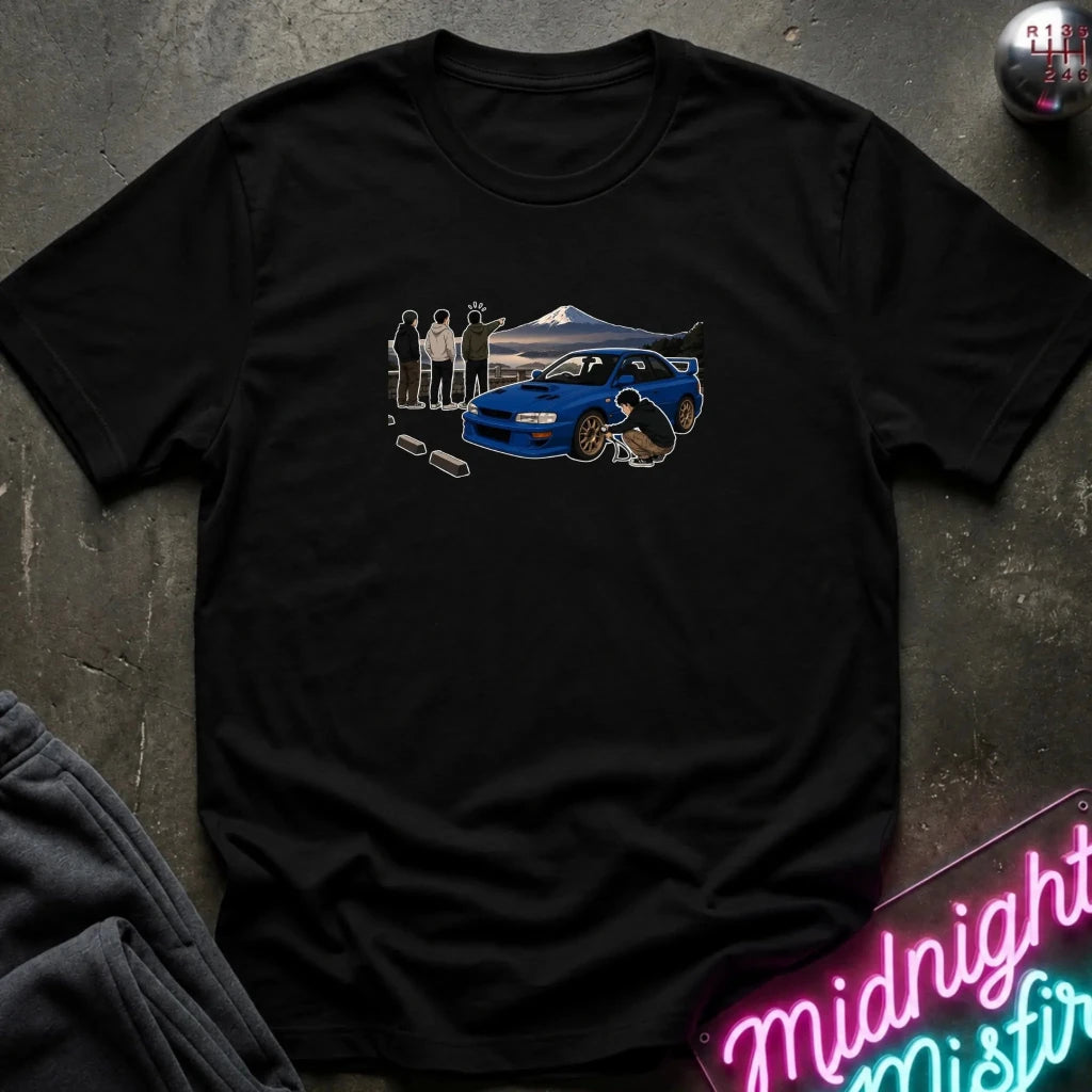 black crewneck t-shirt laid flat on a dark textured surface. The shirt features a graphic print of a blue sports car with a driver and spectators in front of a mountain landscape.