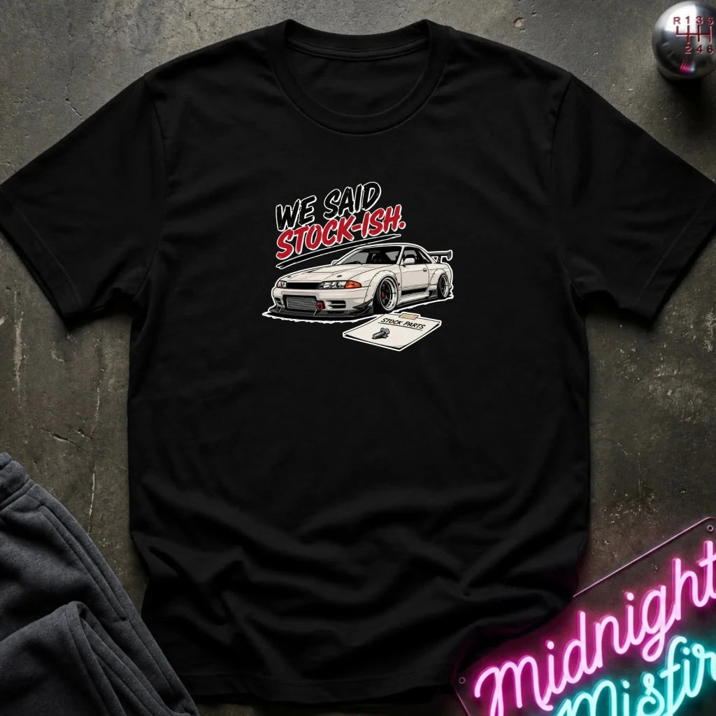A black short-sleeve graphic t-shirt laid flat on a dark textured surface. The graphic features the text 'WE SAY STOCKISH' in white and red, above a white sports car and a small cassette tape illustration.