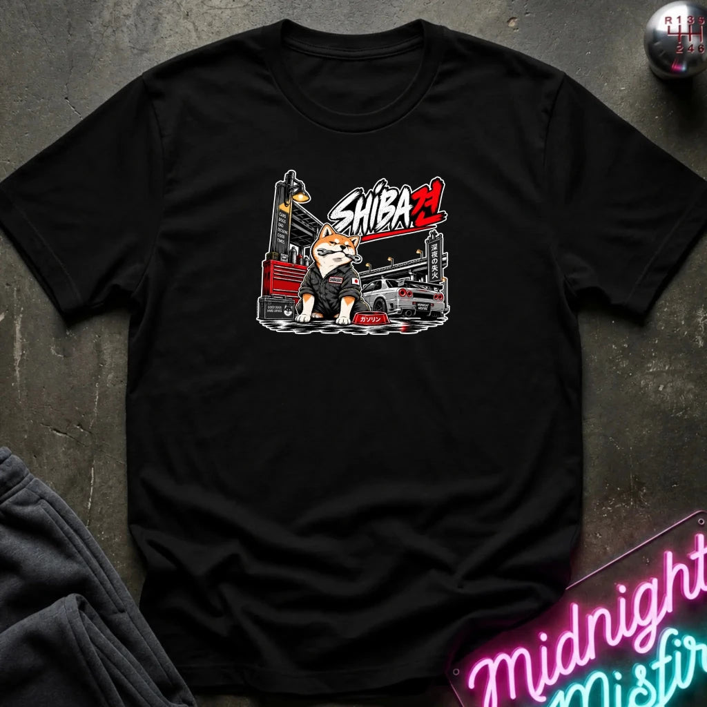 A black short-sleeve graphic T-shirt laid flat on a dark textured surface. The shirt features a central graphic of a Shiba Inu dog character with a tool chest and the text 'SHIBA' in white and red.
