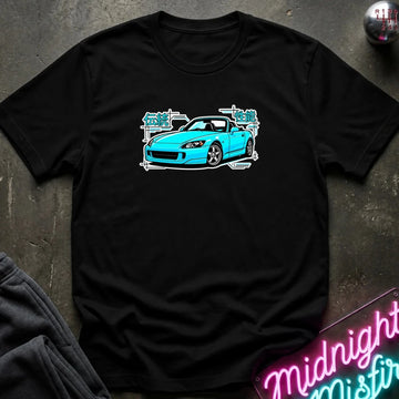 black t-shirt laid flat on a dark textured surface, featuring a graphic print of a bright blue sports car with white and teal text elements. A neon sign and dark clothing are partially visible in the styled flat-lay composition.