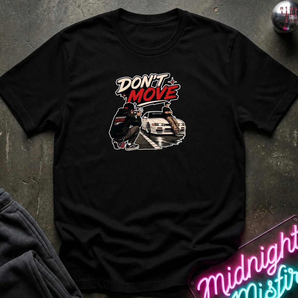 A black short-sleeve graphic t-shirt laid flat on a dark textured surface. The graphic features the text 'DON'T MOVE' in white and red, above an illustration of a person crouching next to a white car.