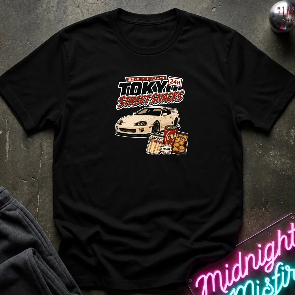 black short-sleeve graphic T-shirt laid flat on a dark textured surface. The shirt features a central graphic with the text 'Tokyo Street Snacks' above a white sports car and snack illustrations.
