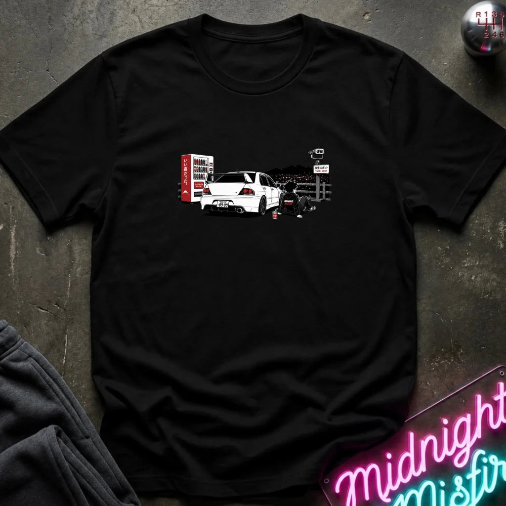 A black short-sleeve t-shirt laid flat on a dark textured surface, featuring a graphic print of a white sports car with a red vending machine.