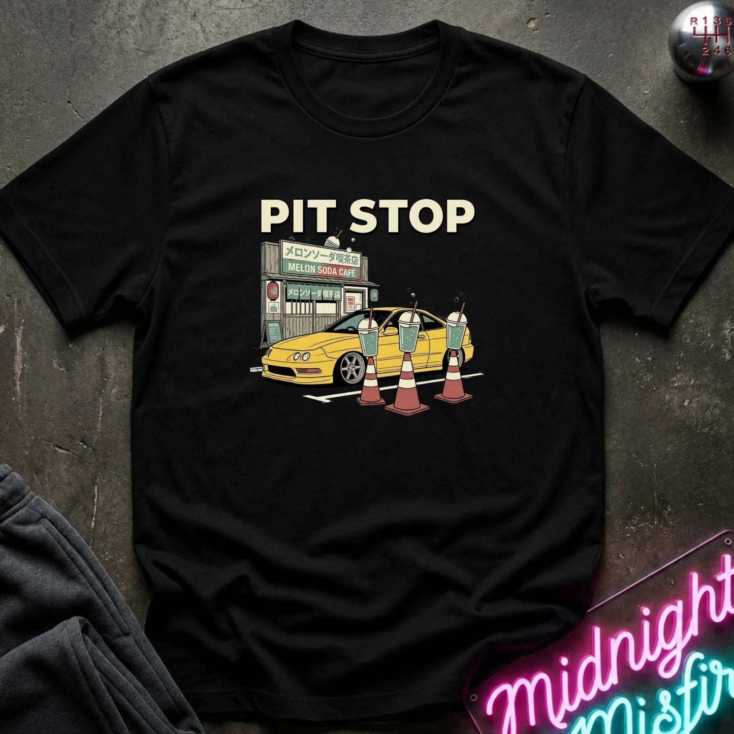 A black short-sleeve graphic t-shirt laid flat on a dark textured surface. The graphic features the text 'PIT STOP' above a yellow car at a cafe with traffic cones.