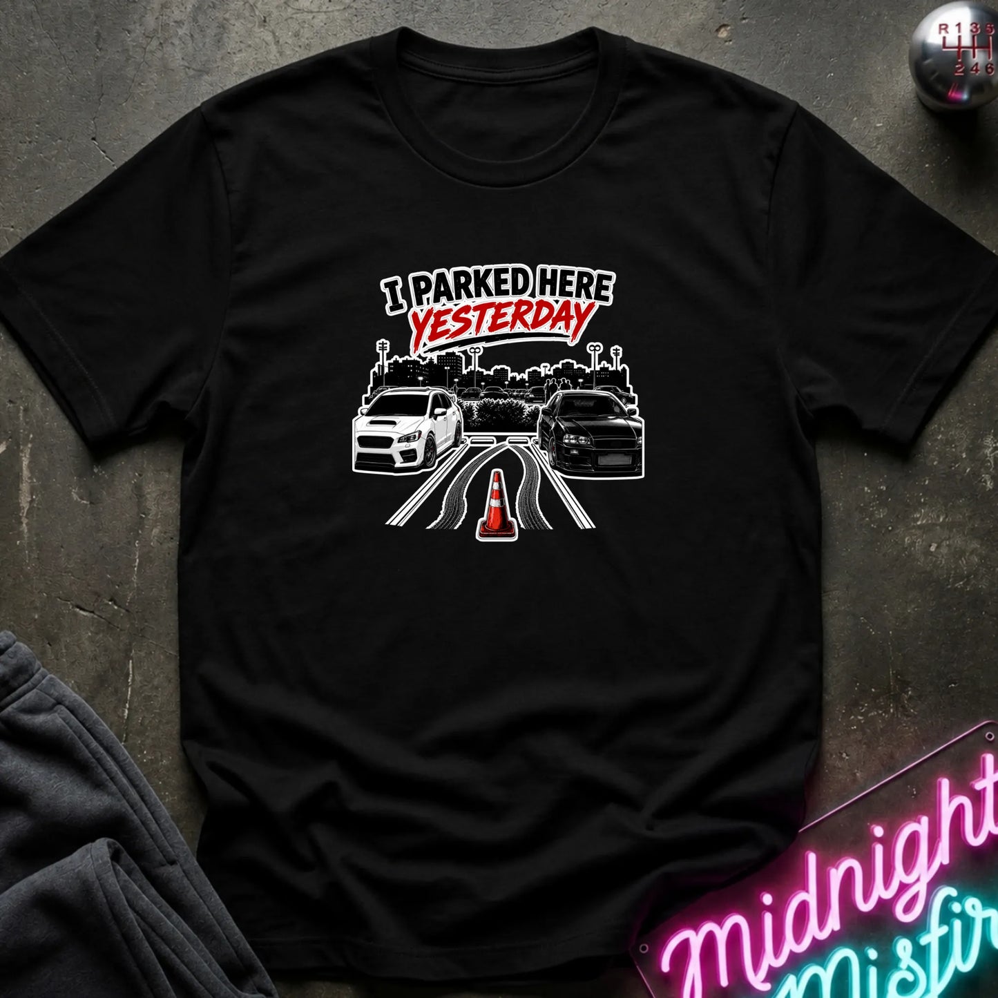 A black short-sleeved t-shirt  that has 2 cars parked in a space in between empty space with skid marks.