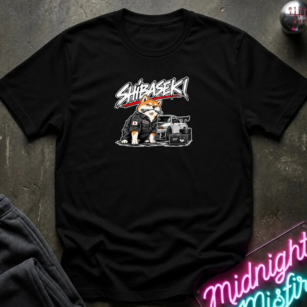 A black short-sleeve graphic t-shirt laid flat on a dark textured surface. The shirt features a graphic of a Shiba Inu dog wearing a black jacket next to a black car, with the word 'SHIBASEKI' written above in a stylized white and red font.
