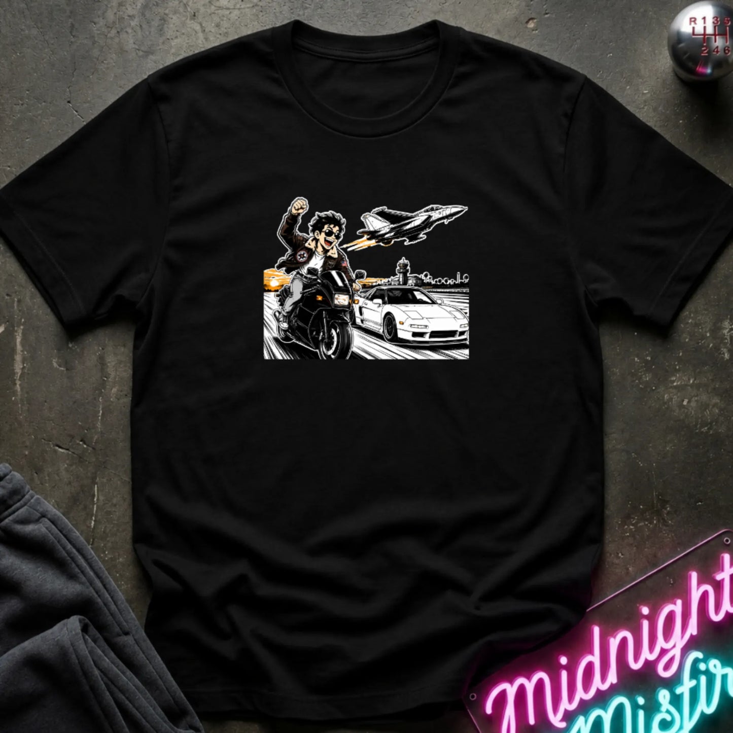 A black short-sleeve graphic t-shirt laid flat on a dark textured surface. The graphic features a white and gray illustration of a woman on a motorcycle next to a white sports car, with a jet plane above.