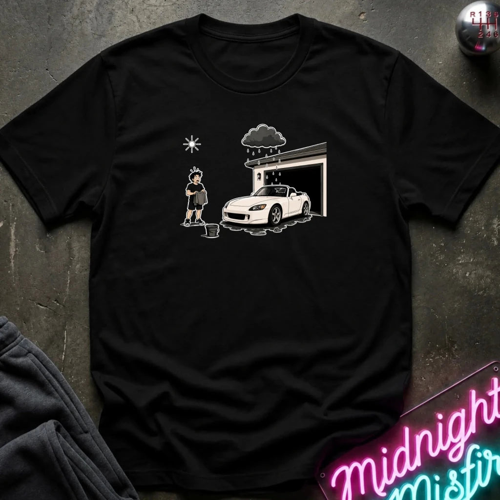 A black short-sleeve graphic t-shirt laid flat on a dark textured surface. The graphic shows a white sports car parked in front of a garage with a person standing nearby.