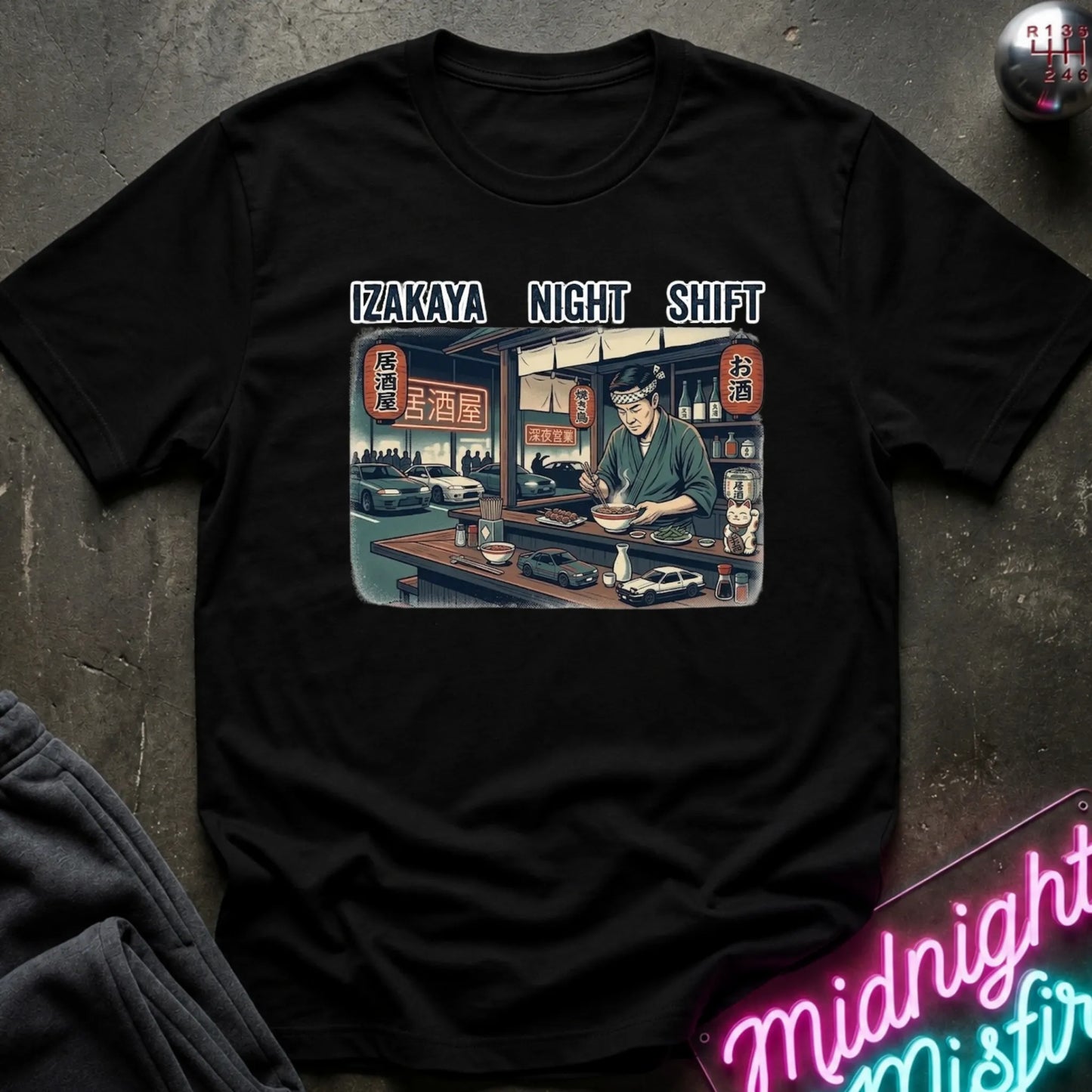 A black short-sleeve graphic t-shirt laid flat on a dark textured surface. The shirt features a rectangular anime-style print of a bartender in a Japanese izakaya restaurant, with the text 'IZAKAYA NIGHT SHIFT' printed above the graphic.